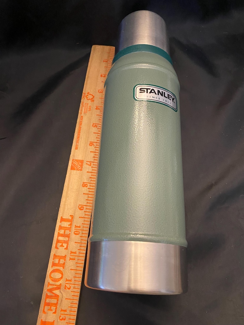 May include: A green Stanley thermos with a silver base and top. The thermos has a label that reads "STANLEY SINCE 1913". A wooden ruler is placed next to the thermos.