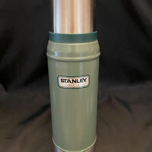 May include: A green Stanley thermos with a silver lid and base. The thermos has a cylindrical shape with a green band near the top. The Stanley logo is displayed on the front. The thermos is designed to keep beverages hot or cold.