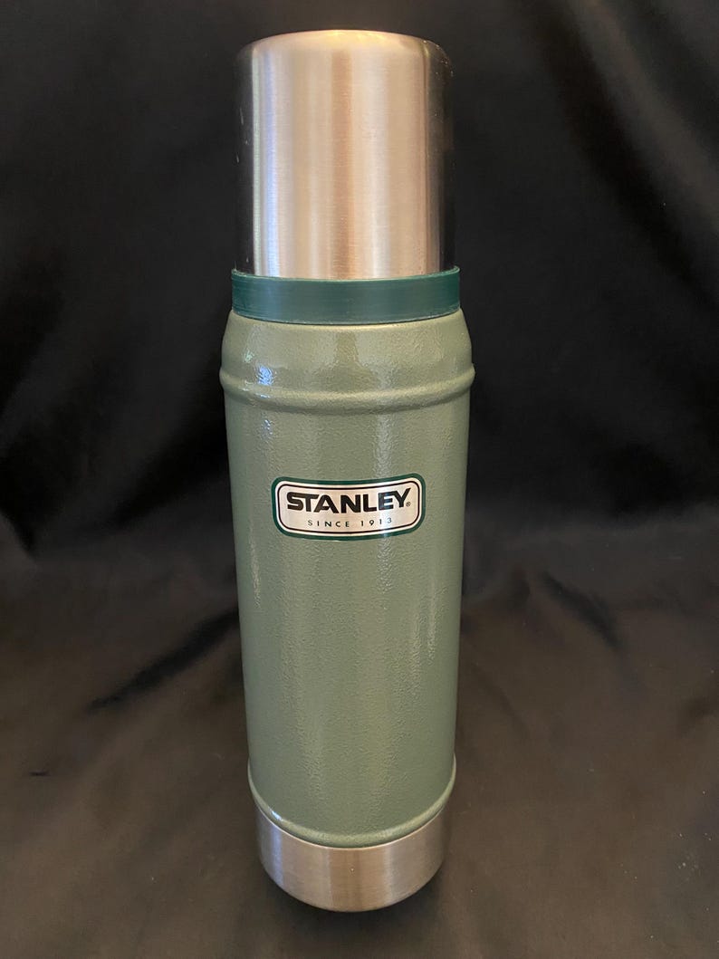 May include: A green Stanley thermos with a silver lid and base. The thermos has a cylindrical shape with a green band near the top. The Stanley logo is displayed on the front. The thermos is designed to keep beverages hot or cold.