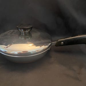 May include: A stainless steel frying pan with a lid. The pan has a black handle with a metal ring. The lid has a black knob. The pan is on a dark surface.