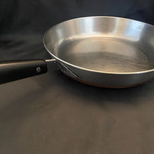 May include: A round metal frying pan with a polished silver interior and a smooth, black handle attached by a single visible screw. The exterior base of the pan features a copper-colored finish, and the overall design is simple and functional.