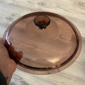 May include: A clear, round, glass lid with a brown knob. The lid has a slightly pinkish hue and a ridged design around the knob. The lid is designed to fit a pot or pan.