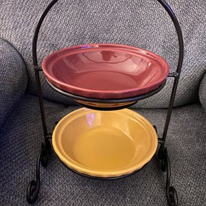 May include: A two-tiered serving stand featuring a black metal frame with a curved handle and scroll-style feet. The stand holds two round ceramic bowls with fluted edges, with the top bowl in a deep maroon color and the bottom bowl in yellow.
