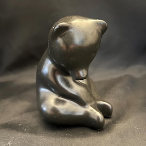 May include: A small, smooth, matte black figurine of a seated bear. The bear is depicted in a hunched, downward-facing posture with its head tilted toward its chest, showcasing rounded ears, a defined snout, and curved limbs in a resting position.