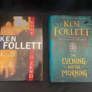 May include: Two hardcover books by Ken Follett are displayed. The book on the left is titled "Code to Zero" with a black, white, and red cover. The book on the right is titled "The Evening and the Morning" with a teal cover.