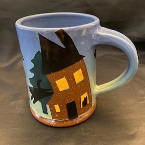May include: A blue ceramic mug with a brown house and a black cat silhouette design. The house has yellow windows and a brown door.