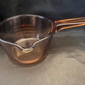 May include: Amber-colored glass measuring cup with a handle. The cup has a pouring spout and orange measurement markings. The handle is rectangular and extends from the side of the cup. The cup is designed for kitchen use.