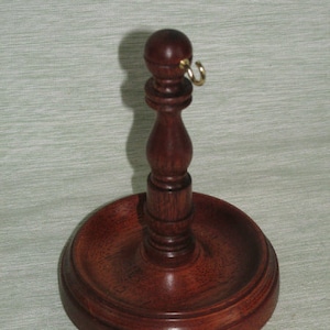 May include: A handcrafted, dark wood pipe stand with a round base and a central stem topped with a small ball and a gold-colored ring. The stand is designed to hold a pipe and is made of polished wood.