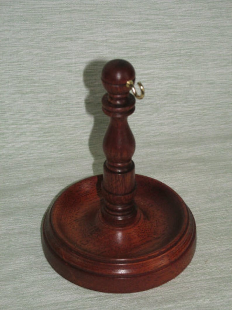 May include: A handcrafted, dark wood pipe stand with a round base and a central stem topped with a small ball and a gold-colored ring. The stand is designed to hold a pipe and is made of polished wood.