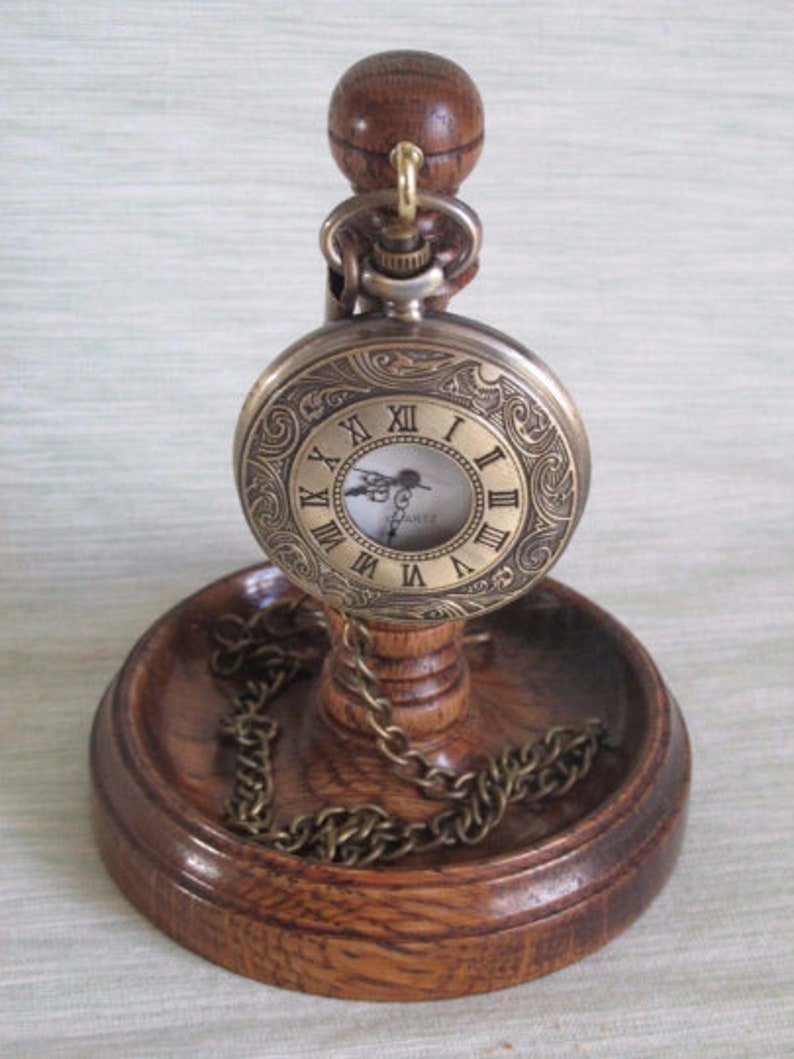 May include: An antique-style pocket watch with a decorative engraved face, Roman numerals, and a chain, displayed on a wooden stand. The stand has a round base and a small wooden ball at the top to hold the watch.