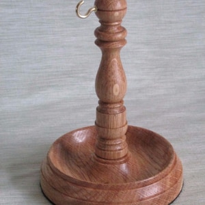 May include: A wooden pipe stand with a round base and a central column. The stand is made of light brown wood and features a brass hook at the top. The base has a recessed area for holding a pipe. The stand is approximately 6 inches tall.