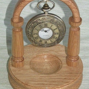 May include: A wooden stand with an arched top for displaying a pocket watch. The stand is made of light brown wood and has a small, round indentation in the base. A gold-toned pocket watch with Roman numeral markings hangs from the arch.