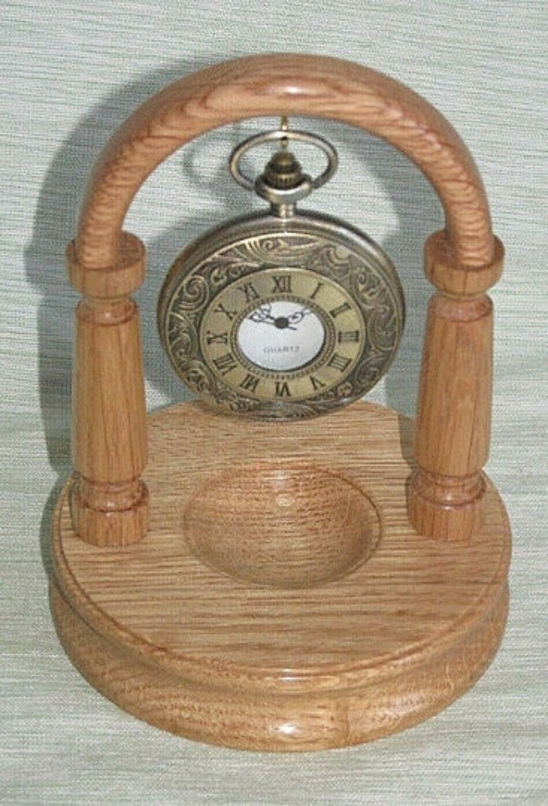 May include: A wooden stand with an arched top for displaying a pocket watch. The stand is made of light brown wood and has a small, round indentation in the base. A gold-toned pocket watch with Roman numeral markings hangs from the arch.