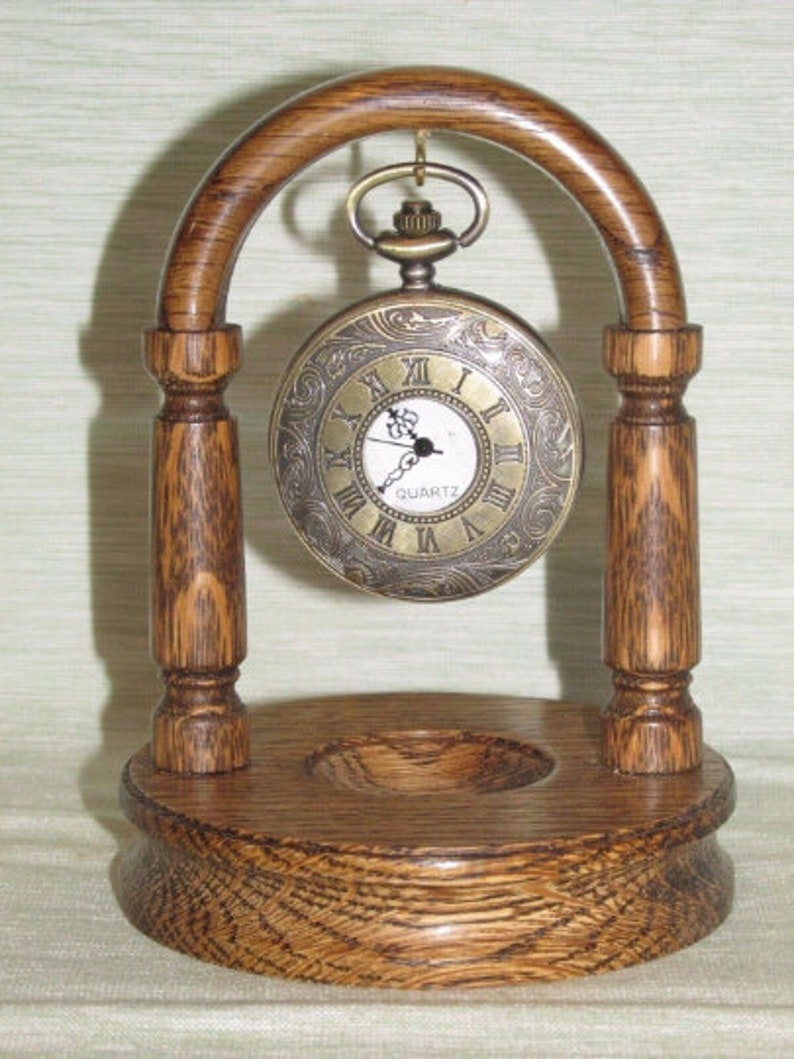 May include: A wooden stand with a curved archway to display a pocket watch. The stand is made of dark wood with a grain pattern. The pocket watch is gold-toned with Roman numeral markings and a white face. The watch face reads "QUARTZ".