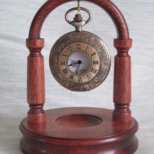 May include: A wooden stand with an arched top holds a gold pocket watch with Roman numeral markings. The stand has a circular base and two pillars.