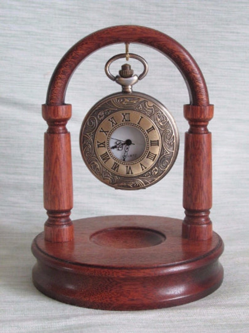 May include: A wooden stand with an arched top holds a gold pocket watch with Roman numeral markings. The stand has a circular base and two pillars.