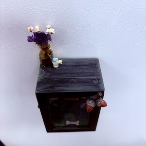 May include: A miniature black wooden box with a glass front, decorated with a small brown vase of purple and white flowers. The box has a miniature scene inside with a bone and a tombstone. Acorn decorations adorn the front.