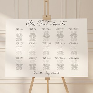 May include: A white seating plan on an easel, featuring the words "Your Seat Awaits" in cursive. The plan lists names under table numbers one to fifteen. The names Isabelle and Craig are listed with the year 2023.