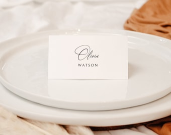 May include: A white place setting with a small rectangular card that reads "Olivia Watson" in elegant script. The card sits on a white plate, with a second plate underneath. The background includes a tan fabric and white linen.