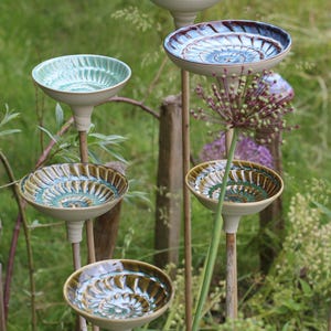 Könnte beinhalten: Eine Ansammlung von fünf Keramik-Gartensteckern, die jeweils eine flache, schalenförmige Oberseite mit einem spiralförmigen, texturierten Muster aufweisen. Die Schalen sind in verschiedenen Farben wie Blau, Grün und Braun glasiert und auf dünnen, hellen Holzstäben montiert.
