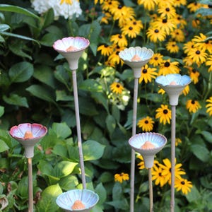Könnte beinhalten: Eine Ansammlung von sechs Keramikblumenskulpturen auf dünnen, hellen Stielen. Jede Blume besitzt eine trichterförmige Blütenstruktur mit einem zentralen, texturierten orangefarbenen Stempel. Die Blütenblätter zeigen Farbverläufe in Rosa, Blau und Weiß.