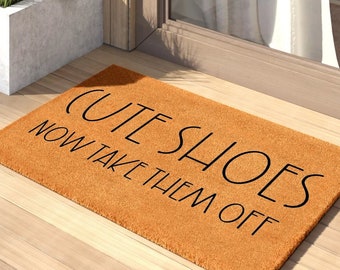 May include: A rectangular doormat in a warm orange hue, featuring the text "CUTE SHOES NOW TAKE THEM OFF" in black, placed on a wooden floor. The mat is positioned near a door, with a planter box visible in the background.