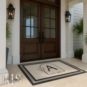 May include: A rectangular doormat with a beige center and a black border is placed in front of a dark brown double door. The mat features a stylized letter "A" with floral accents and the text "EST. 2025".