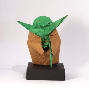 Puede incluir: Una figura de origami verde de un personaje con grandes orejas y una túnica marrón, de pie sobre una base rectangular negra. La figura está hecha de papel plegado, mostrando detalles intrincados y ángulos agudos. El fondo es blanco.