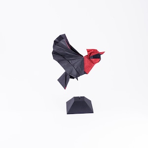 Puede incluir: Una escultura de pájaro de origami negro y rojo, posada sobre una base negra. Las alas del pájaro están extendidas, mostrando los pliegues detallados del papel. La escultura está sobre un fondo blanco.