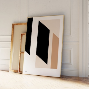 May include: An abstract art print featuring geometric shapes in black, beige, and white. The artwork is framed in gold and leans against a white wall. A second, unframed canvas is visible behind it, adding depth to the composition.