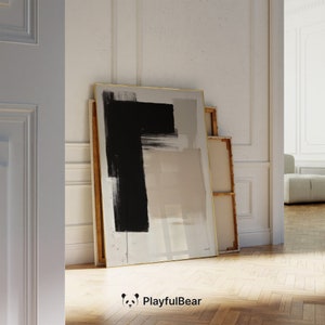 Pode incluir: Impressão de arte abstrata com uma grande pincelada preta sobre um fundo neutro. A obra de arte é emoldurada com uma borda dourada e está encostada a uma parede. A estética é moderna e minimalista.