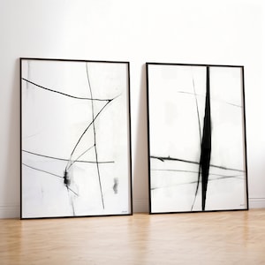 May include: Two framed abstract art prints. Each print features black lines and shapes on a white background. The frames are black. The artwork is displayed on a light wood floor against a white wall.