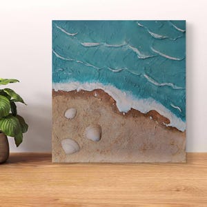 Può includere: Un'opera d'arte su tela quadrata e strutturata che raffigura una scena di spiaggia. L'opera presenta un oceano turchese con onde bianche e spumose, una spiaggia sabbiosa e tre conchiglie. L'arte è esposta su una superficie di legno.