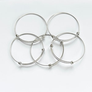 May include: A collection of silver-coloured, adjustable bangle bracelets displayed on a white surface. Each bracelet features a simple, circular design with a hinge and clasp mechanism. The bracelets are made of a shiny, metallic material.