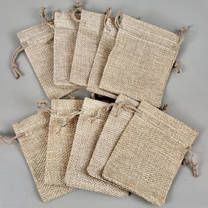 May include: A collection of small, beige burlap drawstring bags. The bags are rectangular with a textured weave and a natural, earthy tone. Each bag has a drawstring closure at the top. The bags are arranged on a grey surface.