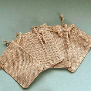 May include: Five small, rectangular burlap drawstring bags are arranged on a light teal surface. The bags are a natural tan colour, with a woven texture. Each bag has a drawstring closure made of a matching tan cord.
