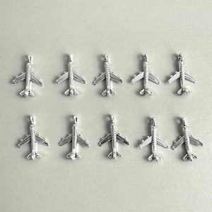 May include: Ten silver-coloured airplane charms arranged on a light grey surface. Each charm features detailed wings, a fuselage, and a loop for attaching to jewellery or other crafts. The charms are evenly spaced.