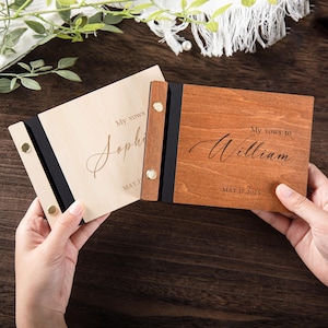Op de afbeelding: Twee houten gelofteboeken met zwarte omslagen en gouden accenten. Het linker boek heeft de tekst "My vows to Sophia" en de datum "MAY 17, 2023". Het rechter boek heeft de tekst "My vows to William" en de datum "MAY 17, 2023".
