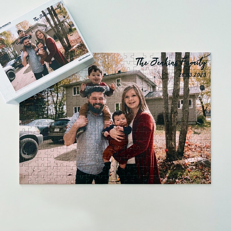May include: A family portrait puzzle featuring a family of four, two adults and two children. The adults are wearing casual clothing and the children are wearing colorful shirts. The puzzle is set against a backdrop of a suburban home and yard. The text "The Jenkins Family 26.3.2023" is printed on the puzzle.