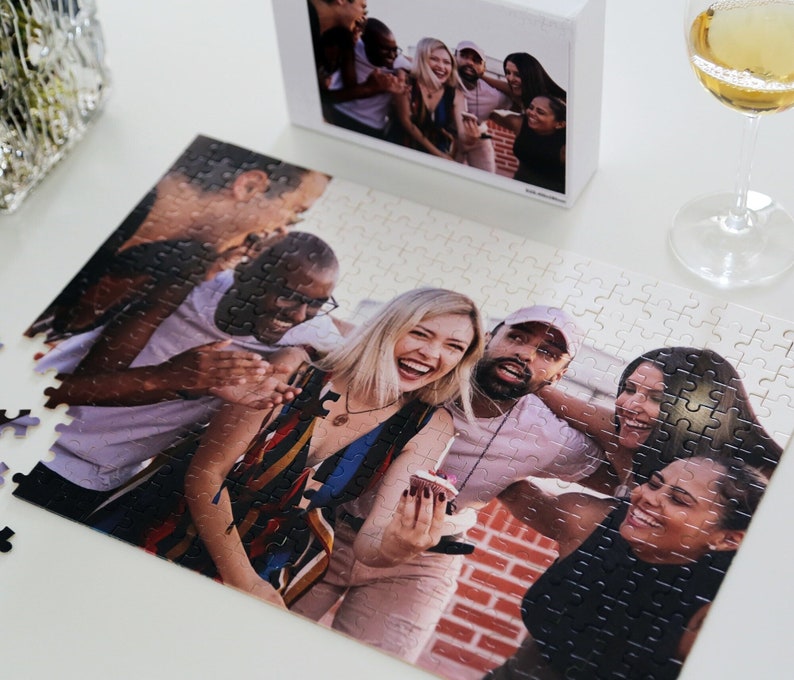 May include: A group of friends pose for a photo while holding a small cake. The friends are smiling and laughing. The photo is printed on a white jigsaw puzzle.