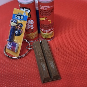 May include: A selection of miniature food-themed items, including two Pringles potato chip containers, a PEZ dispenser, and a Kit Kat candy bar keyring. The PEZ dispenser is blue and yellow, and the Kit Kat is brown.