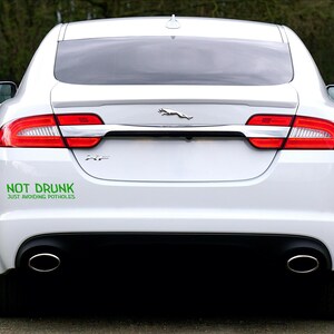 May include: Rear view of a white Jaguar XF car. The vehicle features red rear lights, dual exhaust pipes, and a black lower bumper. A green sticker on the bumper says "NOT DRUNK JUST AVOIDING POTHOLES."
