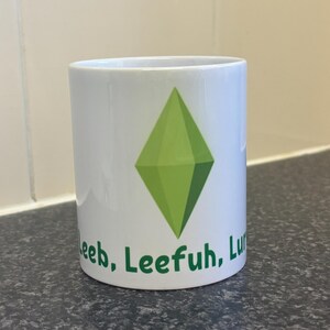 May include: White ceramic mug with a green diamond design and the words "Leeb, Leefuh, Lu" in green. The mug is cylindrical and rests on a dark countertop. The background is a tiled wall.