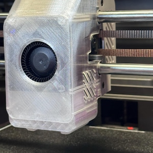 May include: Close-up of a 3D printer's print head assembly. The clear plastic housing encases a fan and other mechanical components. Brown belts and metal rods are also visible, indicating the printer's movement system. The design is industrial and functional.