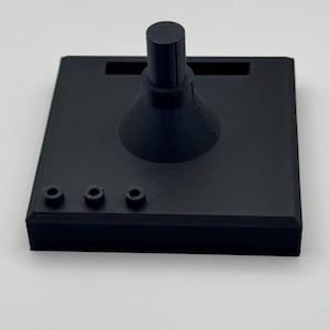 May include: A black 3D-printed stand with a square base. It features a central conical shape topped with a cylindrical post, and three small circular holes on the front edge. The stand is designed to hold an item.