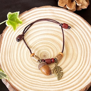 May include: A brown leather bracelet with wooden and red beads, an acorn charm, and a gold leaf charm. The bracelet is displayed on a wooden slice.