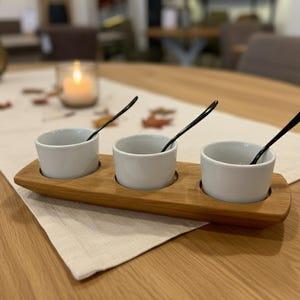 Könnte beinhalten: Ein Holzserviertablett mit drei weißen Keramikschalen, jede mit einem kleinen schwarzen Löffel. Das Tablett besteht aus hellbraunem Holz und liegt auf einem beigen Tischläufer. Im Hintergrund eine brennende Kerze und ein Holztisch.