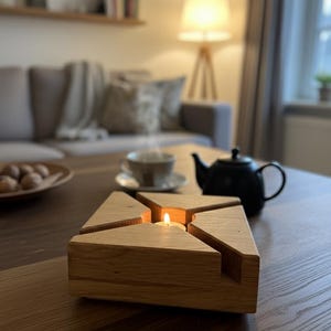 Könnte beinhalten: Ein hölzerner Kerzenhalter mit geometrischem Design. Der Kerzenhalter ist aus hellem Holz gefertigt und hat eine quadratische Form mit dreieckigen Aussparungen. Eine brennende Kerze befindet sich in der Mitte und wirft einen warmen Schein. Eine schwarze Teekanne und Tasse stehen auf dem Tisch.