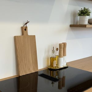 Puede incluir: Una tabla de cortar de madera con asa cuelga de una pared blanca. En una placa de cocina negra, una bandeja dorada contiene dos botellas de aceite de vidrio, dos molinillos de sal y pimienta. Una pequeña planta en maceta está en un estante de madera.