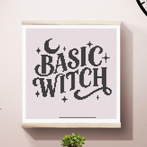 May include: A framed cross-stitch artwork with the words "BASIC WITCH" in black, adorned with stars and a crescent moon. The artwork is displayed on a light pink background and mounted on a wooden frame.
